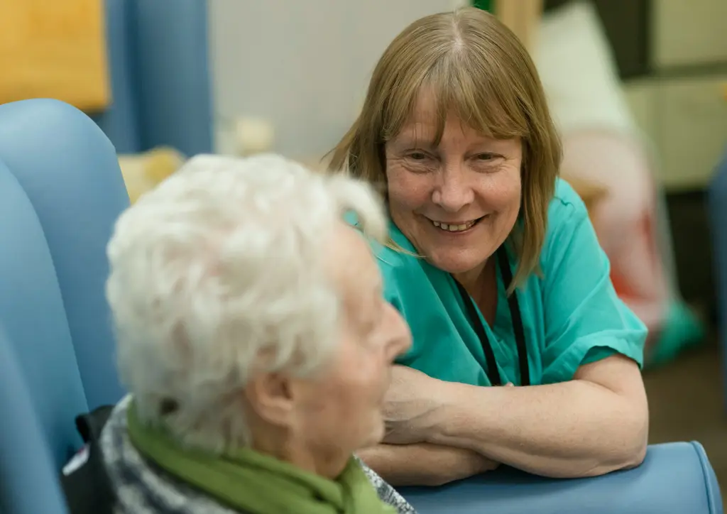 A care worker leans in to talk with an older woman sitting in her chair.