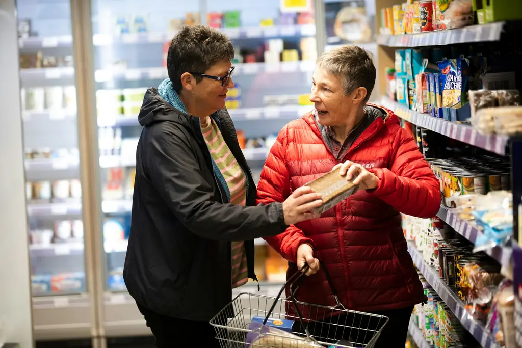 Two friends chat while choosing groceries together in a local shop.