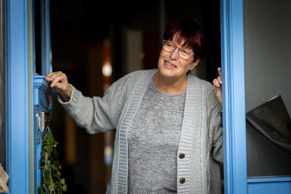 A woman opens her blue front door and smiles at someone arriving.